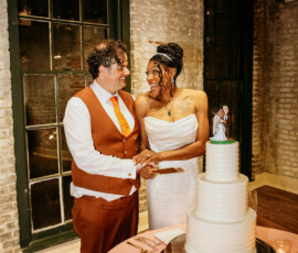 Bride and groom smile as they cut a white wedding cake by candlelight in a brick-walled, warmly lit reception room.