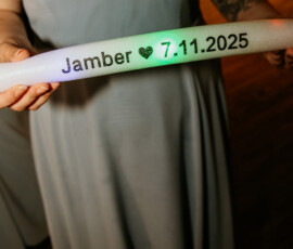 Person in a light gray dress holds a glowing “Jamber ♥ 7.11.2025” foam stick at a lively Louisiana wedding; torso and hands visible.