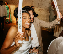 Smiling woman in white dress dances with man in tan suit at energetic Louisiana wedding, holding a drink and Jamber 7.11.2025 light stick.