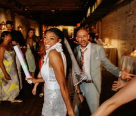 A joyful woman in a sparkling dress and veil dances with friends at a lively Louisiana wedding, surrounded by smiling guests.