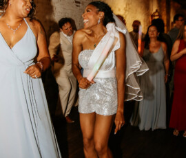 Two women in dresses dance and smile at a Louisiana wedding; one wears a sparkly outfit with a veil and glow stick.