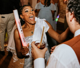 Woman in sparkling dress dances with foam stick at lively Louisiana wedding, smiling at man with drinks; guests in background.