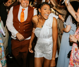 A happy couple in a sparkly outfit and brown suit celebrate at a party, cheered by guests with streamers—wedding inspiration.