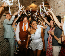 A happy couple walks through cheering guests with streamers in a brick hallway at their Louisiana wedding reception.