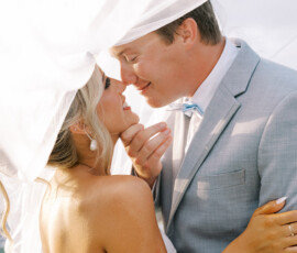 Close-up of a bride and groom embracing under the bride’s flowing veil, smiling as they lean in for a kiss during their wedding portraits.