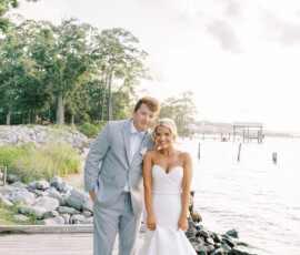 Bride in strapless white gown and boots with groom in light gray suit on dock, Gulf South wedding, water and trees behind.