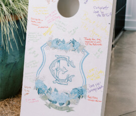 White cornhole board with blue floral C crest and handwritten messages, outdoors by a potted plant at a Louisiana wedding.