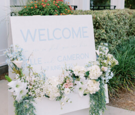 Wedding welcome sign decorated with white and pastel flowers and greenery, displayed outside a venue entrance, welcoming guests to Elle and Cameron’s June celebration.