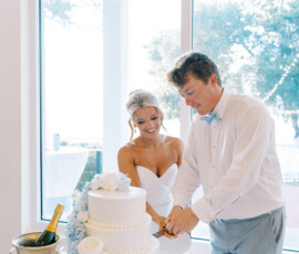 Bride and groom smiling as they cut their white tiered wedding cake decorated with blue and white flowers inside a bright reception venue with large windows.