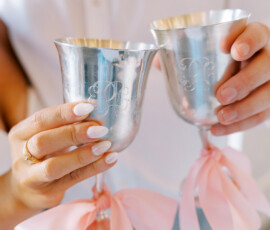 Close-up of a bride and groom holding two silver toasting goblets with monogram engraving and pink ribbon bows tied around the stems.