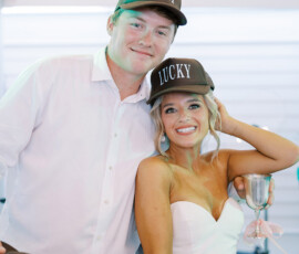 Bride and groom smiling at their wedding reception, both wearing matching hats that say “Lucky,” with the bride holding a silver goblet decorated with a pink ribbon.