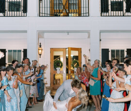 Bride and groom share a kiss as he dips her outside a white Louisiana venue, surrounded by joyful guests in blue.
