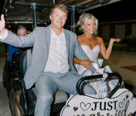 Bride and groom smiling and waving as they ride away in a golf cart decorated with a “Just Married” sign during their nighttime wedding exit.