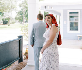 Smiling guest in floral dress and red scarf, with another in a gray suit, outside a light blue house before a New Orleans wedding.