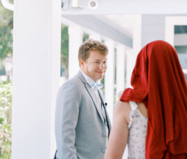 Man in light gray suit smiles at person in floral dress with red headscarf on a bright porch—New Orleans wedding inspiration.