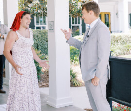 Two men laugh together at a lively Louisiana wedding; one in a suit, the other in a floral dress and red scarf.