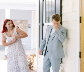 A man in a floral dress and red scarf laughs as he flexes; another in a gray suit smiles by a yellow house at a Louisiana wedding.