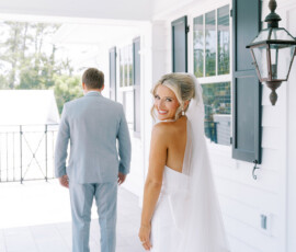 “Bride smiling at the camera before first look with groom in light gray suit on a white porch.”