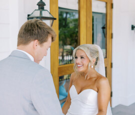 Bride in white dress and veil smiles at groom in light gray suit outside wooden doors, classic New Orleans wedding.