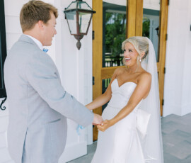 Bride in white dress and groom in light gray suit smile, holding hands outside by wooden doors at a Louisiana wedding.