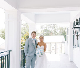 Bride in white gown with bouquet and groom in light gray suit on a sunny, classic New Orleans porch.