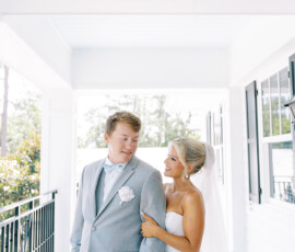 Bride in white dress and veil smiles at groom in gray suit on bright, elegant Louisiana wedding walkway.