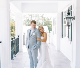 Bride in white gown hugs groom in gray suit on bright porch, capturing their joyful New Orleans wedding before the ceremony.