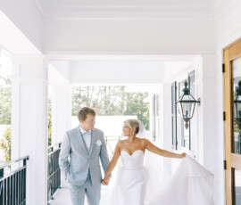 Bride in a strapless white gown and groom in a light gray suit smile, walking hand-in-hand on a bright Louisiana walkway.