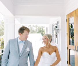 Bride in strapless white gown and groom in light gray suit walk hand-in-hand down a bright hallway, smiling at each other.