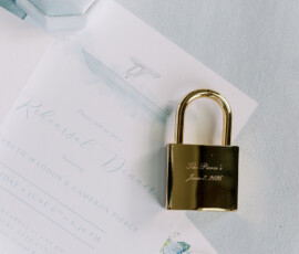 Gold padlock, key, and ring atop light blue wedding stationery with a watercolor rehearsal dinner invitation.