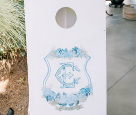 White cornhole board with blue monogram, flowers, and fish motifs on a patio—charming Louisiana wedding inspiration.