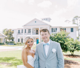 Bride in white dress and groom in light gray suit hold hands, smiling outside a large white building on their sunny Louisiana wedding.