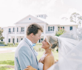 “Bride and groom smiling and embracing outdoors in front of a white Southern-style mansion, with the bride’s long veil flowing in the breeze on their wedding day.”