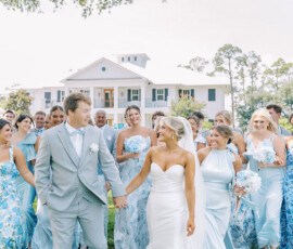 Bride and groom smile, walking hand in hand with their pastel blue bridal party before a white house and trees in Louisiana.