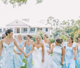 “A smiling bride walks hand-in-hand with her bridesmaids in light blue dresses on a sunny lawn in front of a white Southern-style mansion.”
