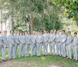 “A large wedding party of groomsmen in light gray suits stands together outdoors under mossy oak trees near the water.”