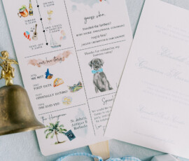 Wedding program fan decorated with illustrations of the couple’s love story, event timeline, and guest travel map, tied with a blue gingham ribbon, pictured next to wedding invitations, rings, and a bell.