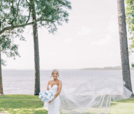 “Bride in a strapless white gown holding a blue and white bouquet with her veil flowing in the breeze by the waterfront.”