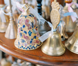 Close-up of a collection of small decorative bells on a wooden display, including a colorful enamel bell and a brass bell with a pineapple handle, each tied with a ribbon.