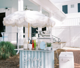 Outdoor snow cone cart at a wedding, featuring a light blue stand with “Snow Cones” lettering, colorful syrups, cups, and a white fringe umbrella in front of a white building.