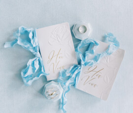“Elegant flatlay of white his and hers wedding vow booklets with blue ribbon, diamond rings, and wedding details on a soft blue background.”