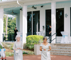 Two elderly women in silver dresses scatter flower petals outside a white building at a sunny Gulf South wedding, camera crew nearby.