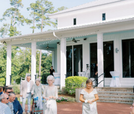 Three older women in light dresses walk an outdoor aisle at a Louisiana wedding, white building and guests in the background.