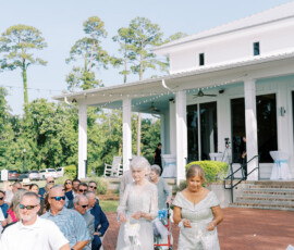 Two elderly women in formal dresses walk down an outdoor aisle at a Louisiana wedding, guests seated, white building behind.
