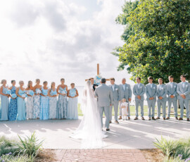 Bride and groom at outdoor altar with blue-clad bridesmaids, gray-suited groomsmen, greenery, and large tree in background.
