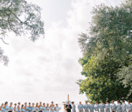 Outdoor New Orleans wedding with bride, groom, blue-clad bridesmaids, gray-suited groomsmen, and guests on white chairs.