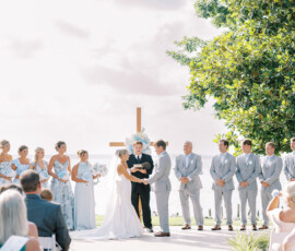 Bride and groom exchange vows outdoors by a wooden cross with bridal party in blue and gray; guests watch elegant ceremony.