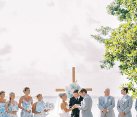 Bride and groom exchanging rings during an outdoor waterfront wedding ceremony, standing beneath a wooden cross decorated with blue and white flowers, surrounded by bridesmaids in light blue dresses and groomsmen in gray suits.
