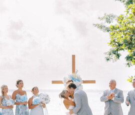 Bride and groom kiss by the water at an outdoor Louisiana wedding with bridesmaids in blue and groomsmen in gray.