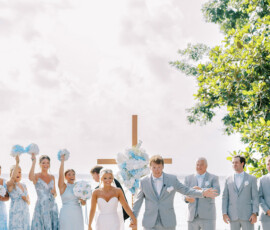 Newlywed couple holding hands and smiling at an outdoor Louisiana wedding with wedding party in light blue and floral cross backdrop.